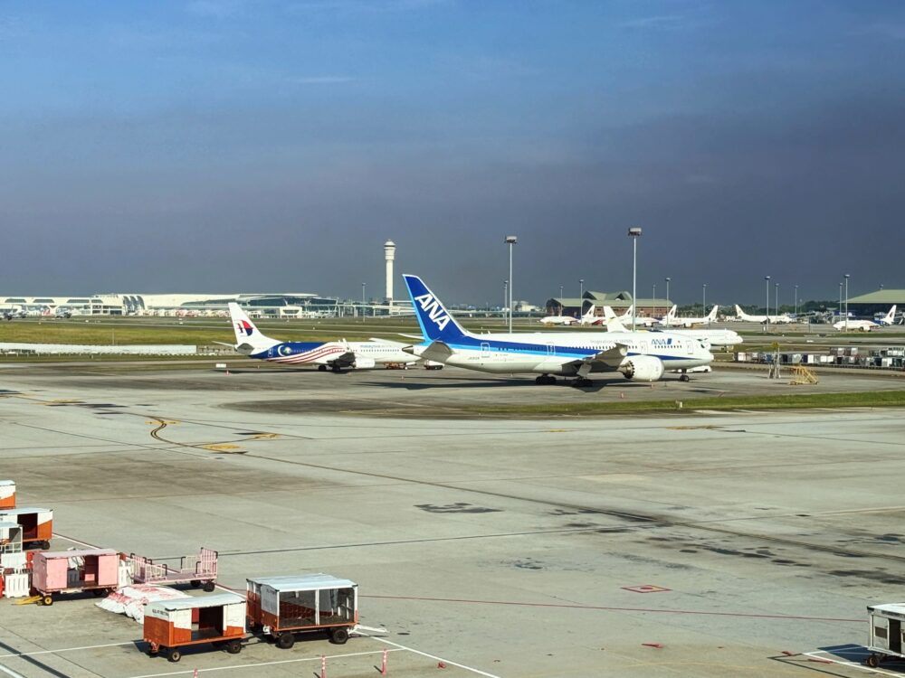 ラウンジの窓から見えるクアラルンプール国際空港のエプロン。手前にカートやULD（航空コンテナ）が置かれ、奥にはANA（全日本空輸）の青い尾翼を持つボーイング機と、マレーシア航空機が駐機している。