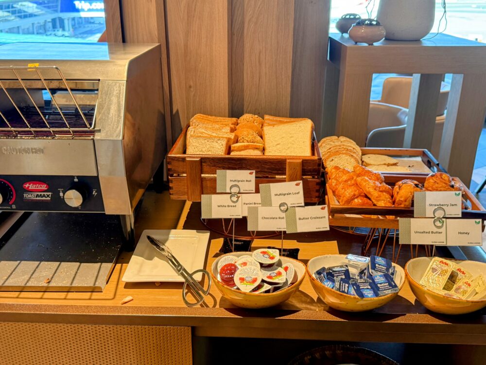 「GLOBAL LOUNGE」のパン・ブレックファーストコーナー。コンベア式トースターの横に、マルチグレインロール、食パン、クロワッサンなどのパンが木製トレイに並び、手前にはバターやジャムのポーションが器に入れられている。