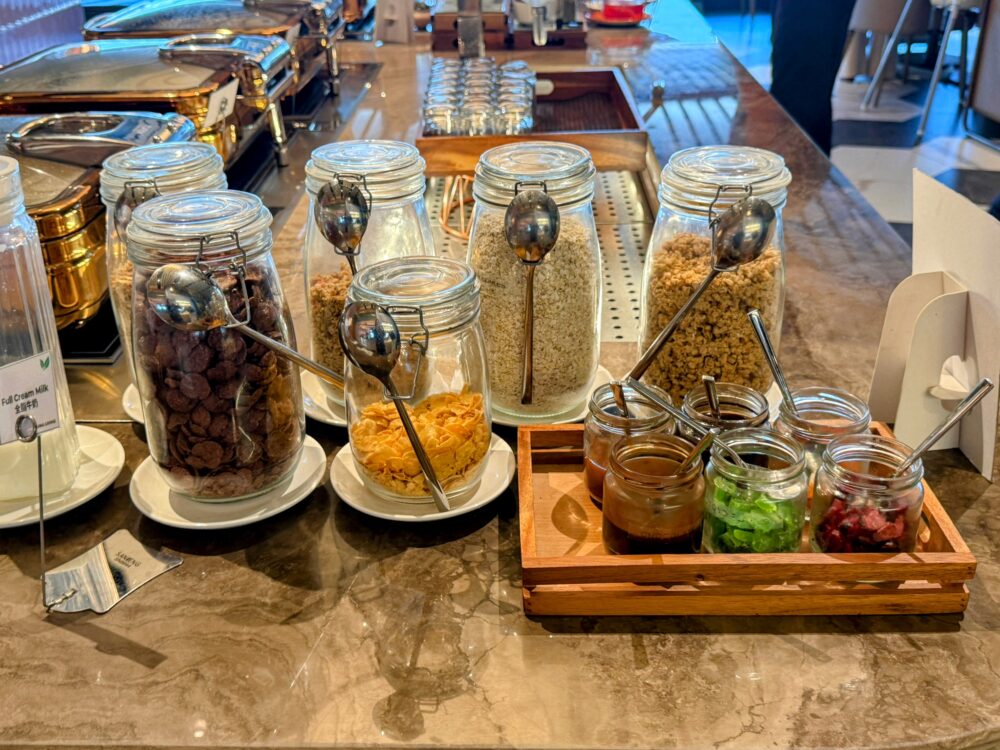 「GLOBAL LOUNGE」のシリアルとトッピングのコーナー。ガラス瓶に入った各種シリアルやオートミールと、チョコレートソース、トッピング用のドライフルーツやゼリーが木製トレイに用意されている。