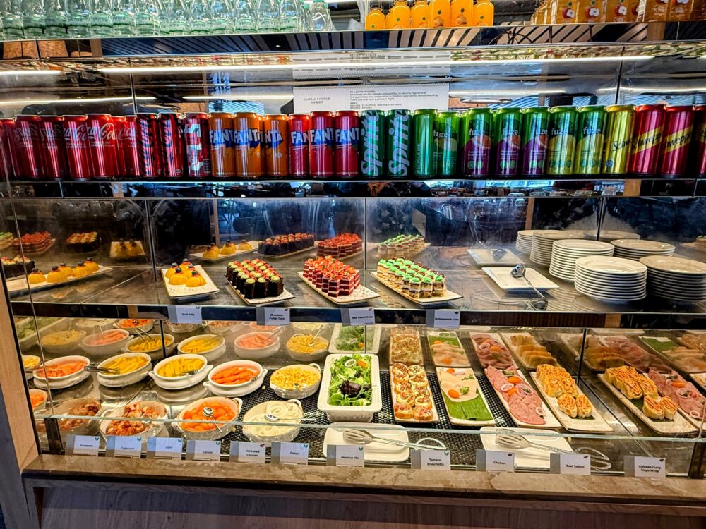 「GLOBAL LOUNGE」の冷蔵ショーケースとビュッフェカウンター。上段には色とりどりの缶ジュースやペットボトルが整然と並び、中段にはカットケーキやデザート類、下段にはサラダやハムなどの冷製料理が並んでいる。