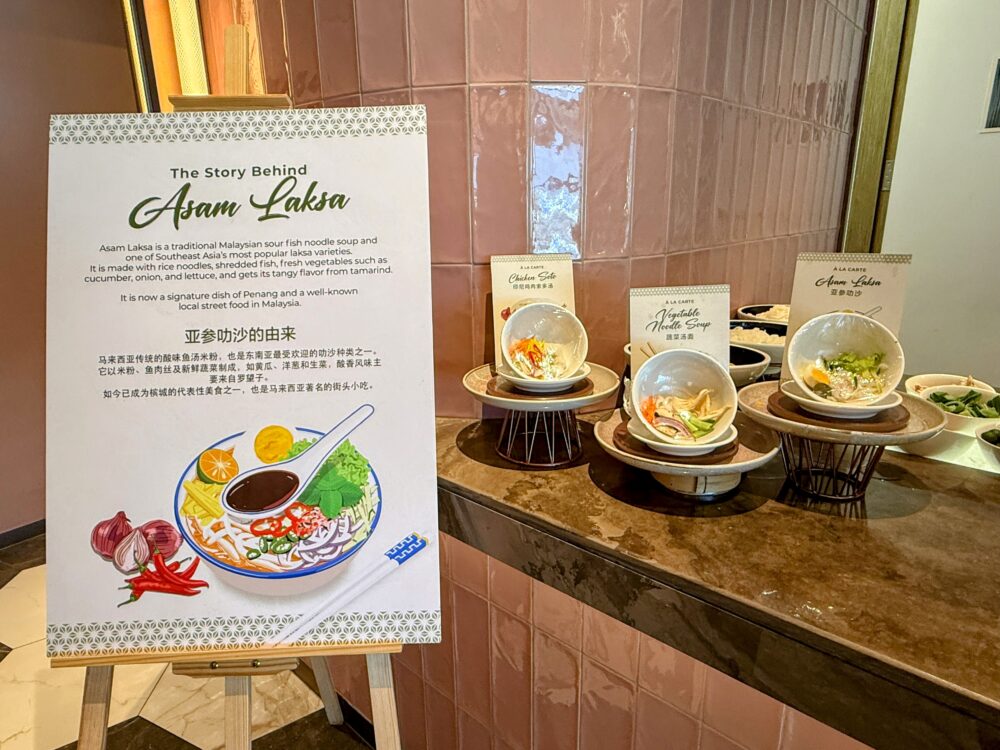 「GLOBAL LOUNGE」のアッサムラクサ（Asam Laksa）の案内看板とヌードルバーのカウンター。名物料理であるアッサムラクサの解説パネルの横に、調理見本のお椀や具材のボウルが配置されている。