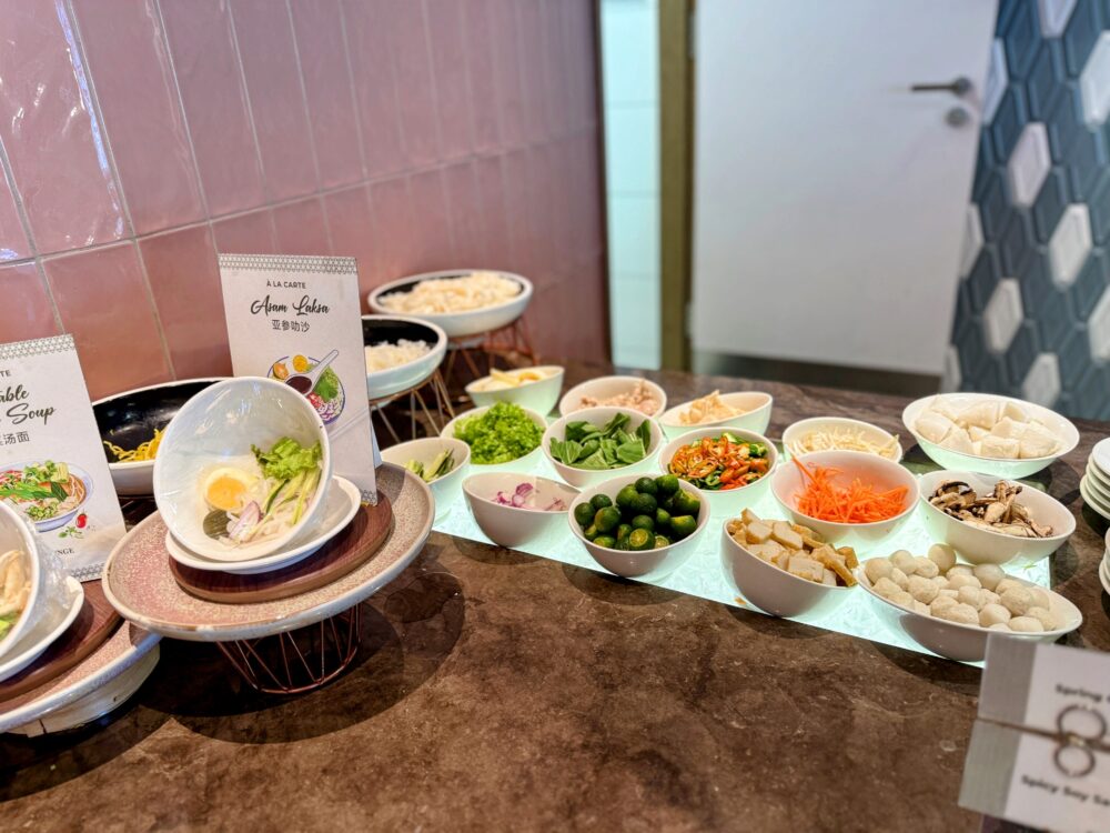 アッサムラクサのトッピングバー。麺が入った器の周りに、細かく刻まれた野菜、ハーブ、ライム、唐辛子、もやし、人参、練り物などの豊富な具材を入れた小さなボウルが冷却カウンターの上に並んでいる。