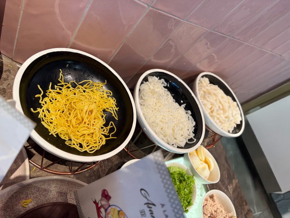アッサムラクサの麺選びコーナーのアップ。黒い縁の白い器に入れられた黄色い中華麺（ホッケンミー）や、白い米粉の麺、フォーのような平打ち麺が種類別に用意されている。