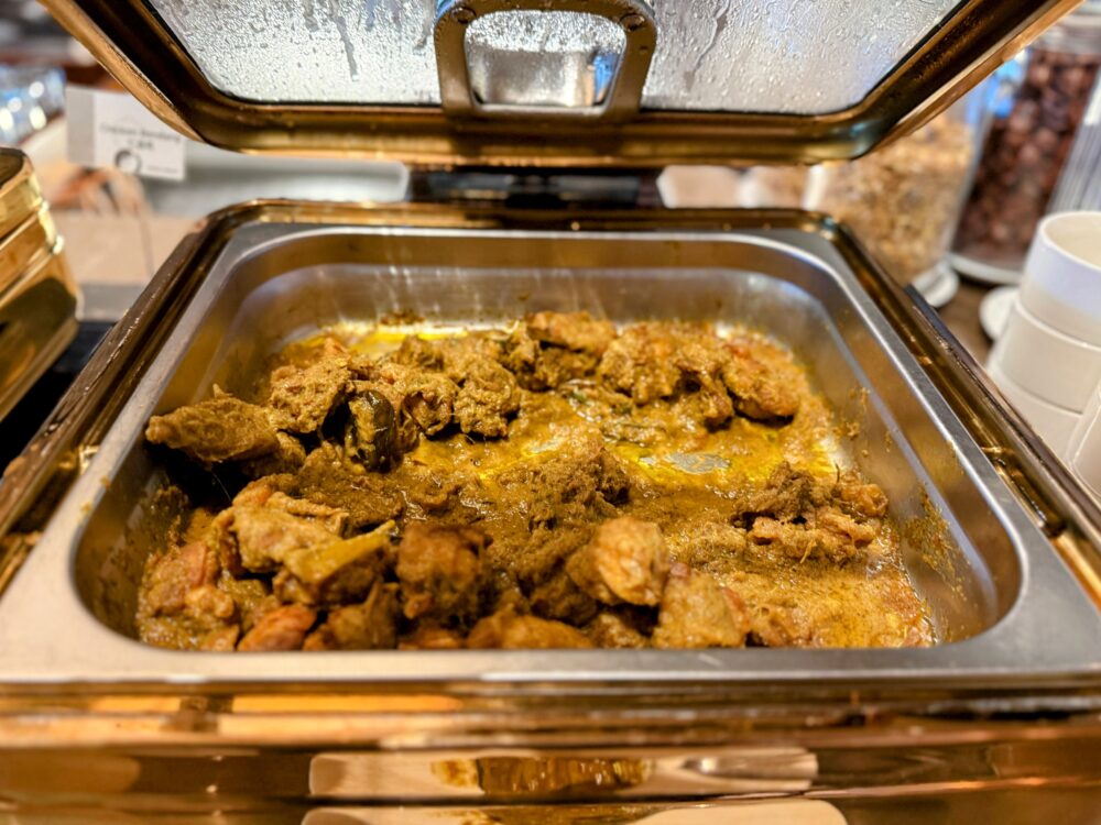 ホットビュッフェのカレー料理（チキンの煮込み）。金色の保温トレイの中に、スパイスとハーブがしっかり効いたチキンと濃厚なグレイビーソースが入っている。