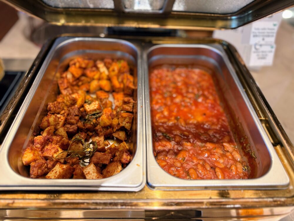 温かいサイドディッシュのホットビュッフェ。左側にはスパイスで味付けされたポテトや豆腐の炒め物、右側にはトマトソースで煮込まれたベイクドビーンズ（豆料理）が入っている。