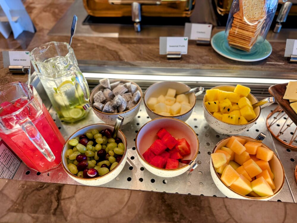 「GLOBAL LOUNGE」のフレッシュフルーツとドリンクのコーナー。デトックスウォーターと真っ赤なジュースのピッチャーの横に、ドラゴンフルーツ、メロン、スイカ、ぶどうなどの色鮮やかなカットフルーツがボウルに盛られている。