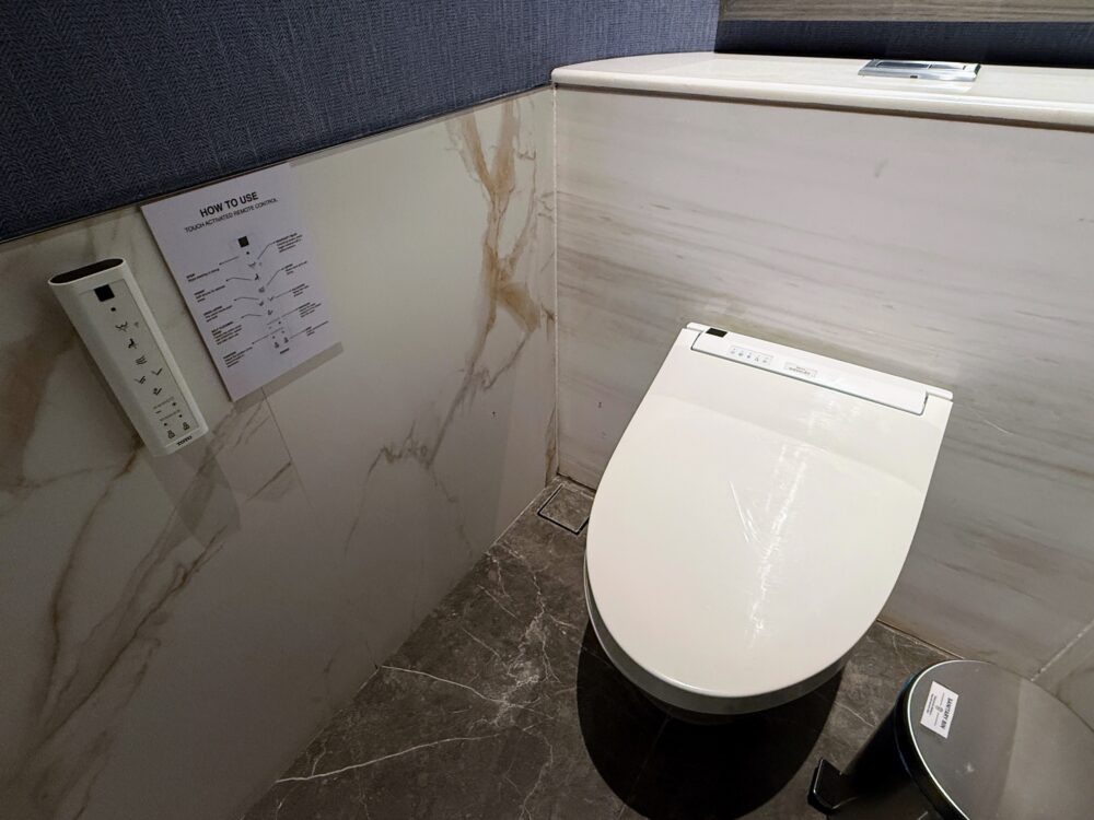 ラウンジ内の洗練された個室トイレの様子。大理石調の壁とダークなタイルの床で統一された空間に、最新式のウォシュレット付きトイレとサニタリーボックスが設置されている。