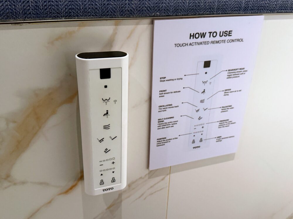 トイレの壁面に設置されたTOTO製のタッチ式リモコンのアップ。洗浄や乾燥などの各種操作ボタンが分かりやすく配置され、隣には英語で使い方を解説した説明シートが貼られている。