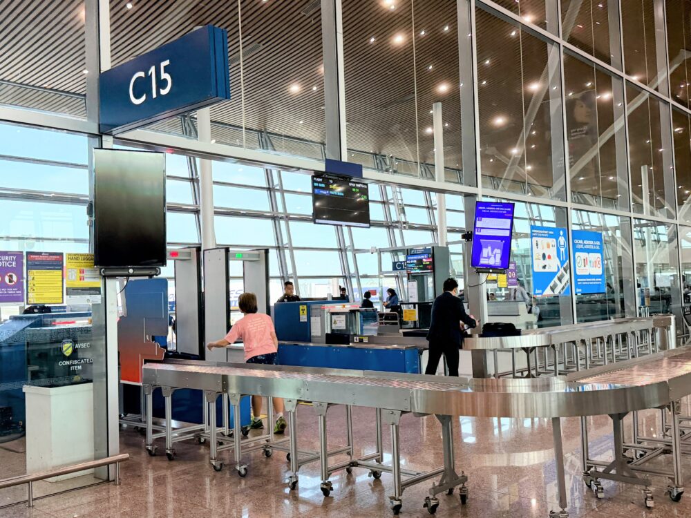 クアラルンプール国際空港（KLIA）サテライトターミナルのセキュリティチェックカウンター（C15ゲート付近）。手荷物のX線検査レーンや金属探知機ゲートが設置され、係員や乗客の姿が見える。
