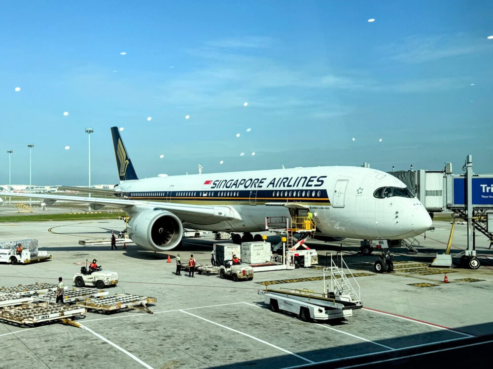 KLIAサテライトターミナルの窓から見える駐機中のシンガポール航空（Singapore Airlines）のエアバスA350機。機体周辺では、トーイングトラクターやULD（航空コンテナ）を運ぶ車両などのグランドハンドリング作業が行われている。