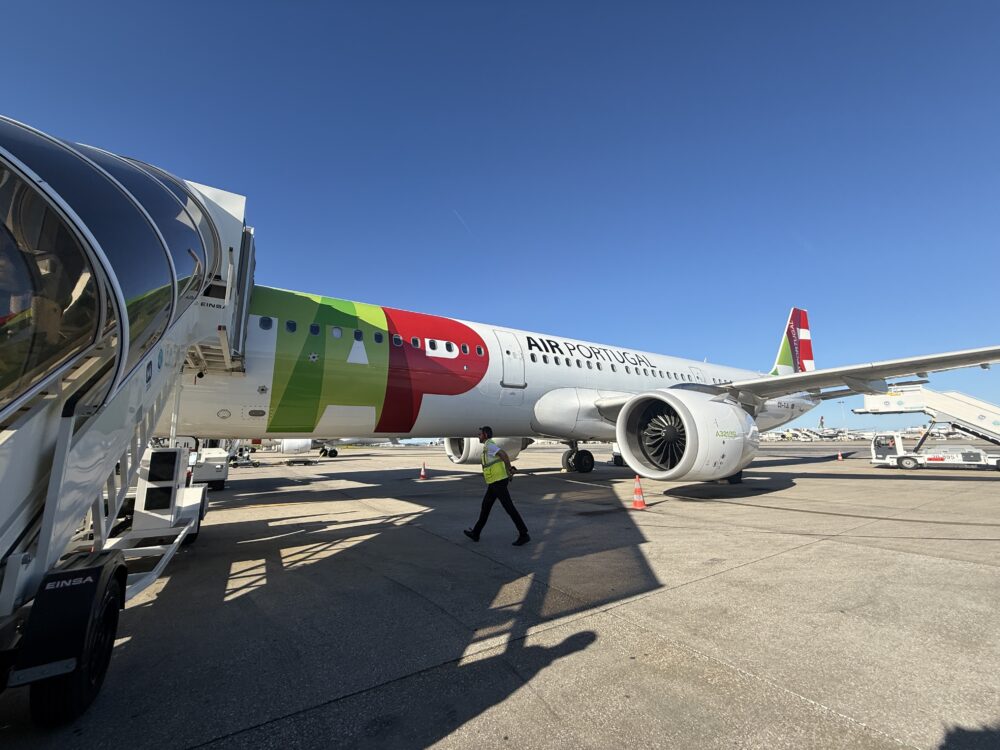 晴れた日の屋外、リスボン空港の駐機場に駐機中のTAPポルトガル航空のエアバスA320型機。胴体には大きな「TAP」のロゴと「AIR PORTUGAL」の文字。手前にはガラス張りのボーディングブリッジが伸び、黄色の反射ベストを着た地上職員が歩いている。他の地上支援車両や遠くの滑走路も見える。