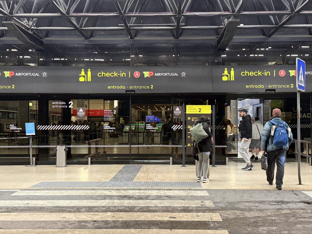 屋内、天井の黒いトラス構造の下にあるTAPポルトガル航空の「check-in(チェックイン)」カウンターの入口。黄色のサインで「entrada - entrance 2」と書かれている。ガラス張りの自動ドアの向こうにチェックインホールが見える。手前には横断歩道のペイントがあり、乗客が歩いており、リュックサックを背負った人がカウンターに向かっている。