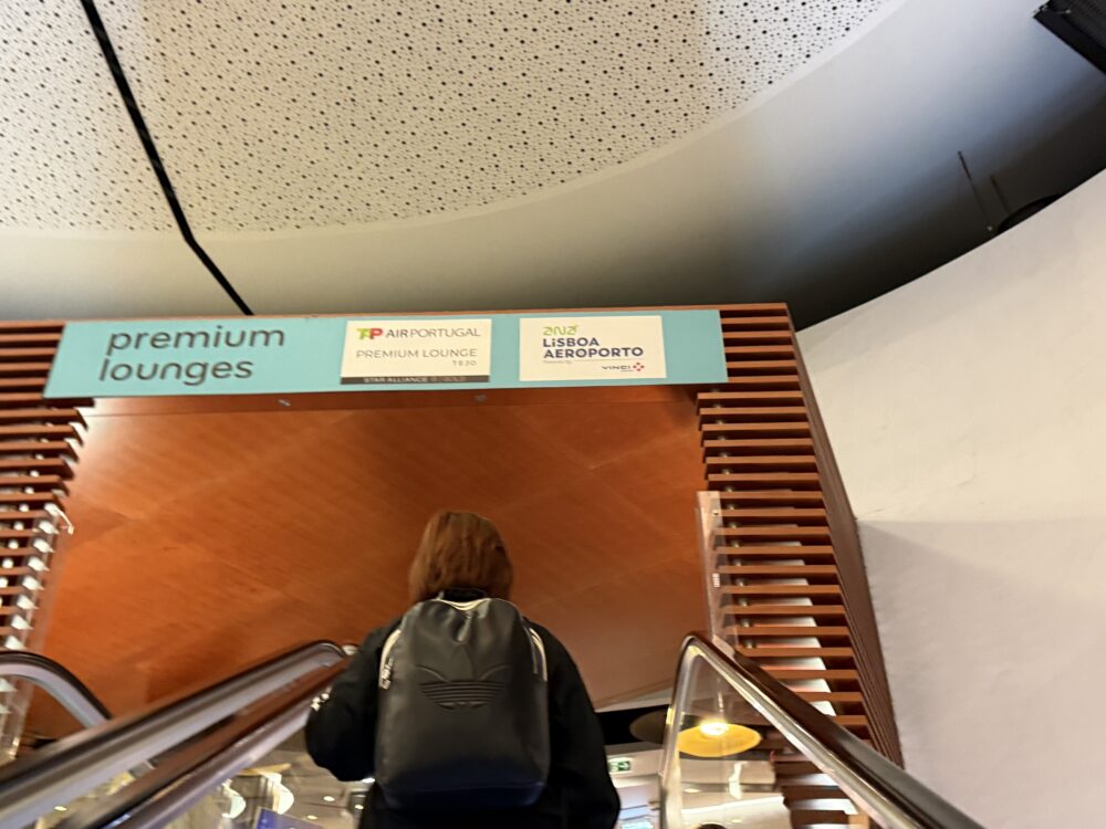2階へ昇るエスカレーターからの視点。頭上には木製の天井とルーバーに囲まれた「premium lounges」の入口サインがある。サインには「TP AIR PORTUGAL PREMIUM LOUNGE TEJO」と「ANA LISBOA AEROPORTO」のロゴと文字が記載されている。エスカレーターに乗っている人物の背面が写っており、アディダスのロゴが入った黒いリュックサックを背負っている。