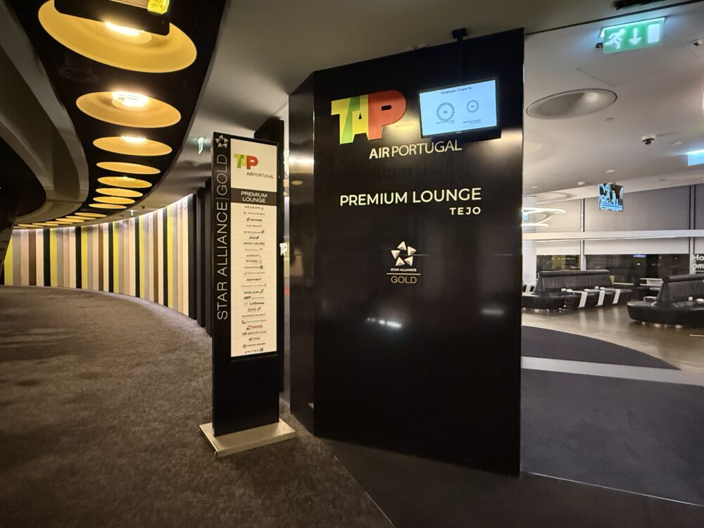黒を基調としたTAPプレミアムラウンジの入口。看板には「TAP AIR PORTUGAL PREMIUM LOUNGE TEJO」やスターアライアンスゴールドのロゴ、提携航空会社の一覧が掲示され、上部には現在の混雑状況を示すモニターが設置されている。