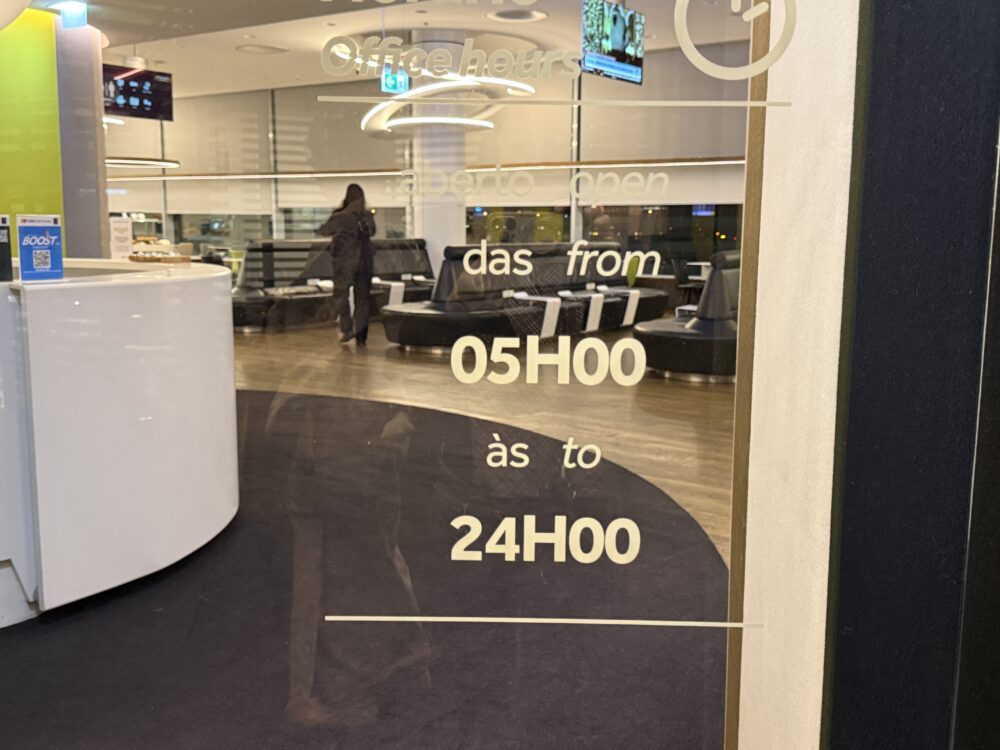 ラウンジのガラス自動ドアに貼られた営業時間を示す文字。「Office hours / aberto open」として「das from 05H00 / às to 24H00」の時間が大きく記載されており、奥のレセプションエリアがうっすらと透けて見えている。