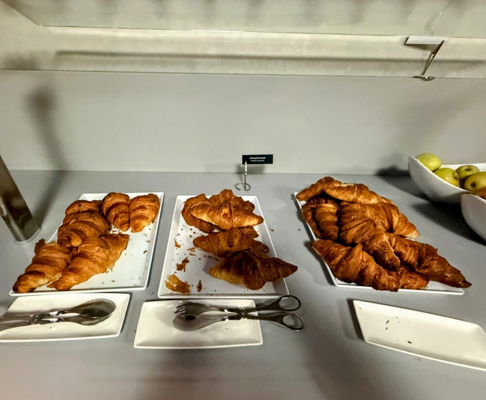 ビュッフェカウンターに並ぶ種類の異なるクロワッサンの数々。「Croissant Francês(フレンチクロワッサン)」の小さな黒い名札が立てられ、きれいに焼き上げられたクロワッサンが3つの白い大皿に山盛りになっている。右奥のボウルには黄色や緑色のリンゴなどのフルーツが見える。