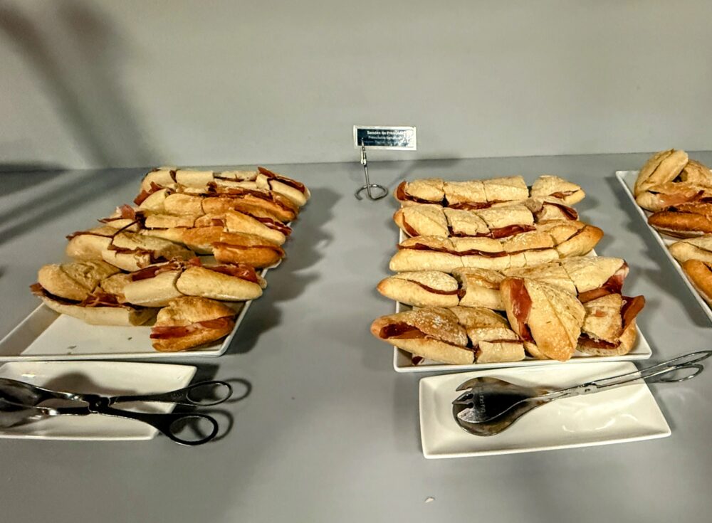 ビュッフェカウンターに並ぶ軽食のサンドイッチ。「Sandes de Presunto(生ハムのサンドイッチ)」と書かれた名札の前に、バゲットに生ハム(プロシュート)が挟まれたサンドイッチが白い皿に整然と並べられている。手前にはトングが置かれている。