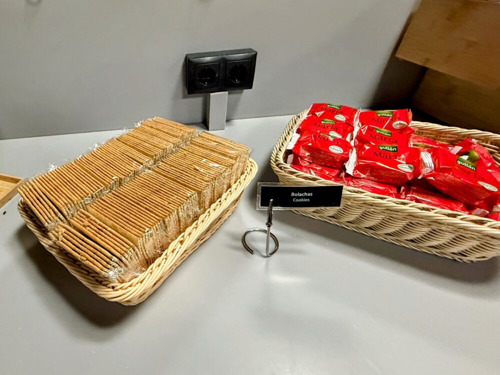 ビュッフェカウンター上のスナックコーナー。「Bolachas / Cookies」と書かれた名札が立てられ、左側のカゴには個包装されていない細長いビスケットが並び、右側のカゴには赤いパッケージに包まれたクッキーが山積みになっている。背後には黒いコンセントプラグが配置されている。