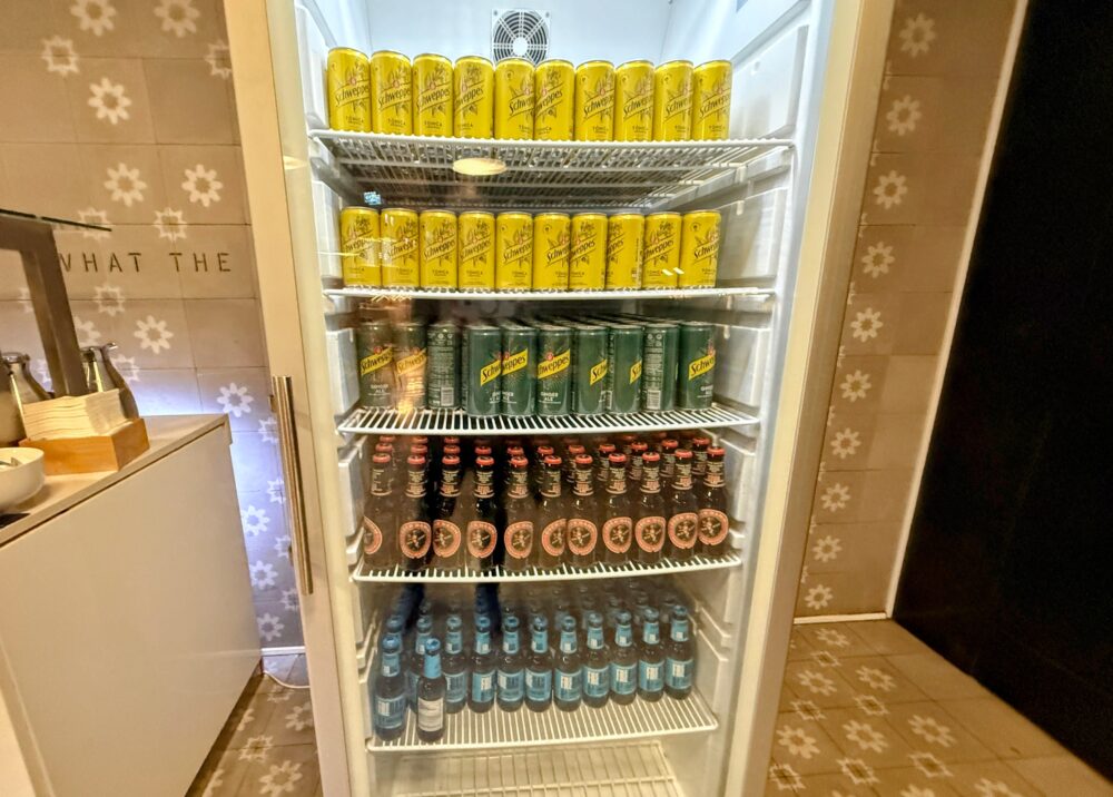 ラウンジ内のドリンク用大型ガラス冷蔵庫。上段から中段にかけて「Schweppes(シュウェップス)」のトニックウォーターやジンジャエールなどの黄色や緑色の缶が整然と並び、下段には茶色の小瓶に入ったビール類や青いラベルのドリンクが冷やされている。