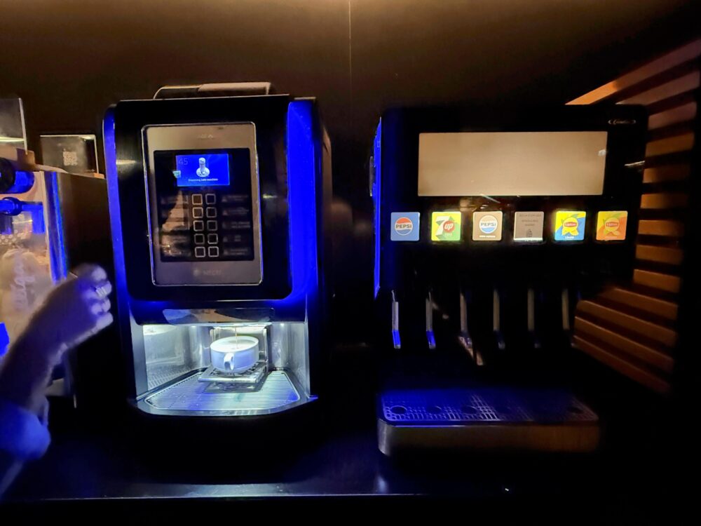 ラウンジ内のドリンクディスペンサーコーナー。左側には青いLED照明に照らされたタッチパネル式の全自動コーヒーマシンが置かれ、カップがセットされている。右側にはペプシや7up、リプトンなどのボタンが並ぶソフトドリンクのサーバー機が配置されている。