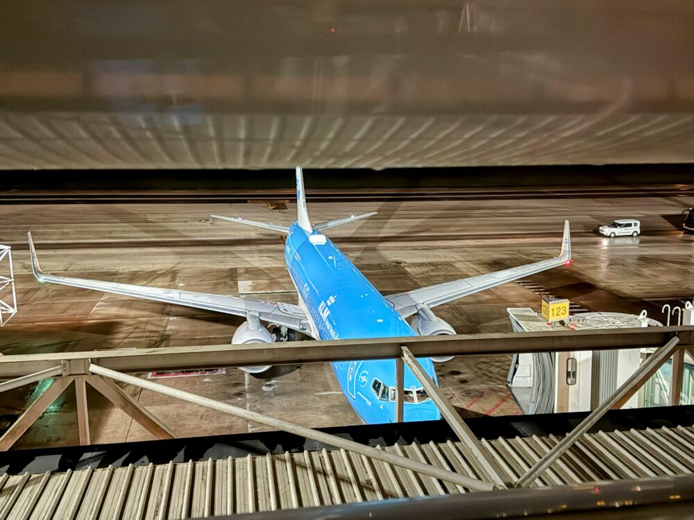 ラウンジの窓越しから撮影した夜間の空港駐機場。手前のボーディングブリッジと構造物の下側に、鮮やかなスカイブルーの塗装が施されたKLMオランダ航空のボーイング737型機が駐機しており、機体後方や主翼のライトが点灯している。