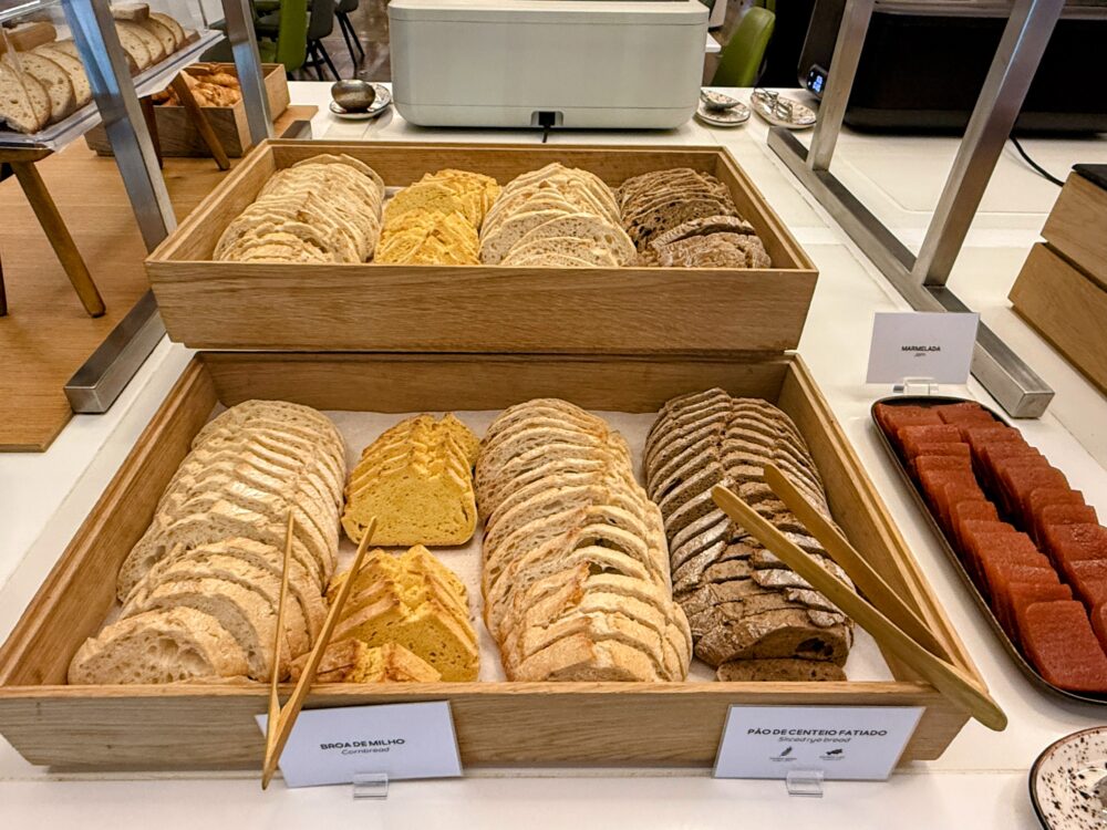 ウッドテイストのビュッフェ台。左側の木箱にはスライスされたコーンブレッド(Broa de Milho)やライ麦パン(Pão de Centeio)がきれいに並び、右側の黒い皿には「Marmelada」と呼ばれるポルトガルの伝統的なマルメロのペースト(羊羹状のスイーツ)がスライスされて並んでいる。