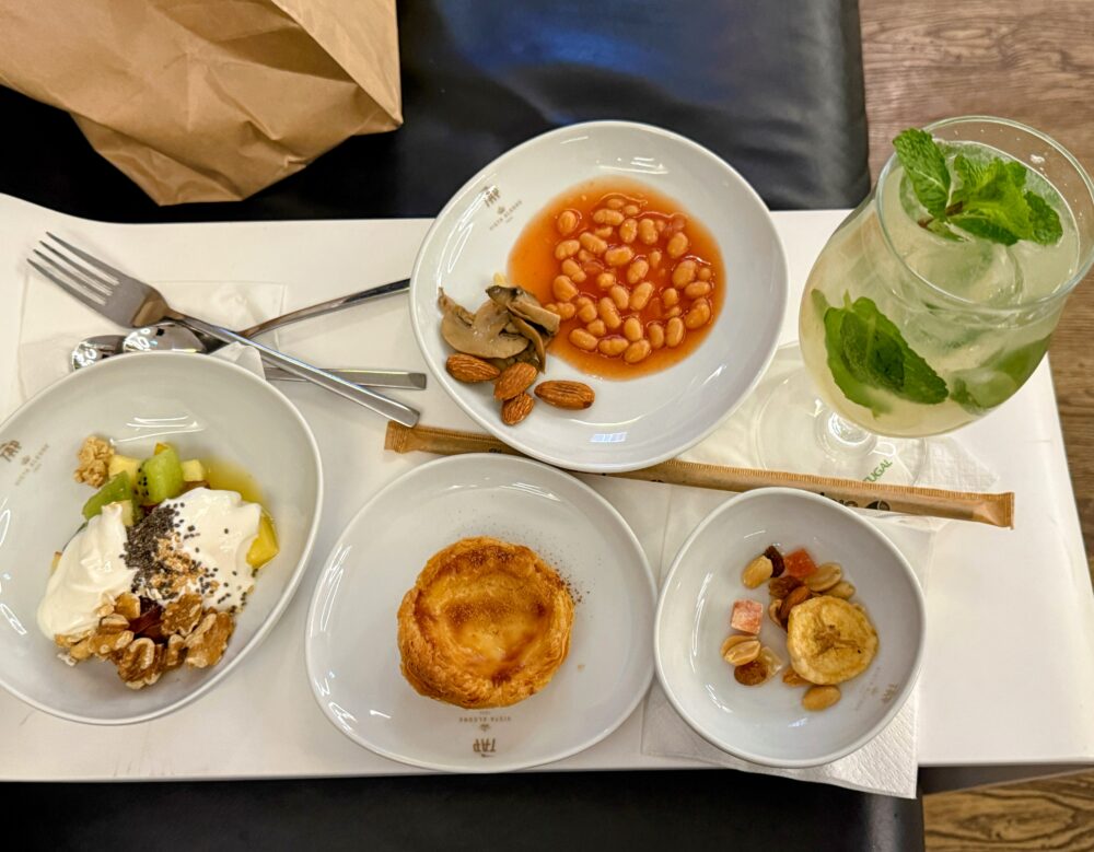 トレイの上に用意された朝食や軽食の数々。ヨーグルト、ベイクドビーンズ、エッグタルト、スナック類がそれぞれの皿に盛られ、右側にはミントの葉が添えられた爽やかなグリーンのカクテルグラスが置かれている。