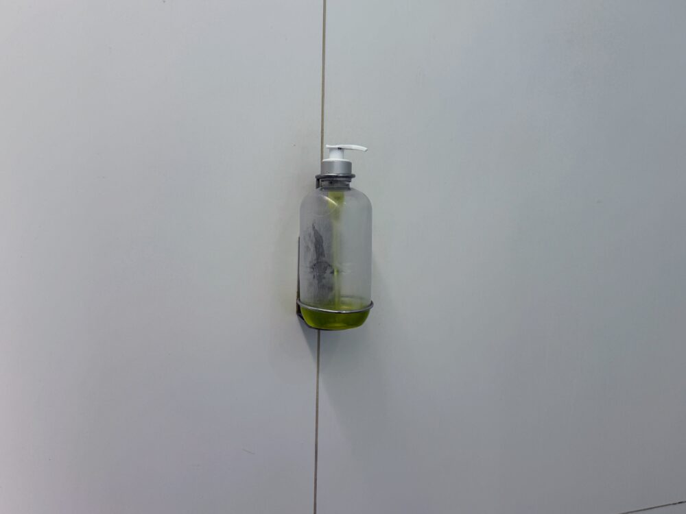 白い壁面に直付けされた透明なボトル型のハンドソープディスペンサー。中には緑色の液体石鹸が入っており、金属製のホルダーにしっかりと固定されている。