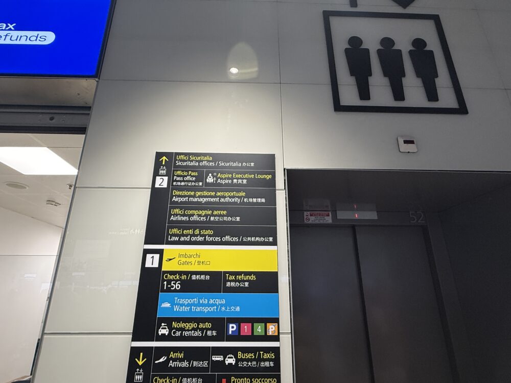 ベネチア・マルコ・ポーロ国際空港の案内標識。「Aspire Executive Lounge」や各種施設、出国ゲート(Gates)の方向を示す表示板とエレベーターの入口。