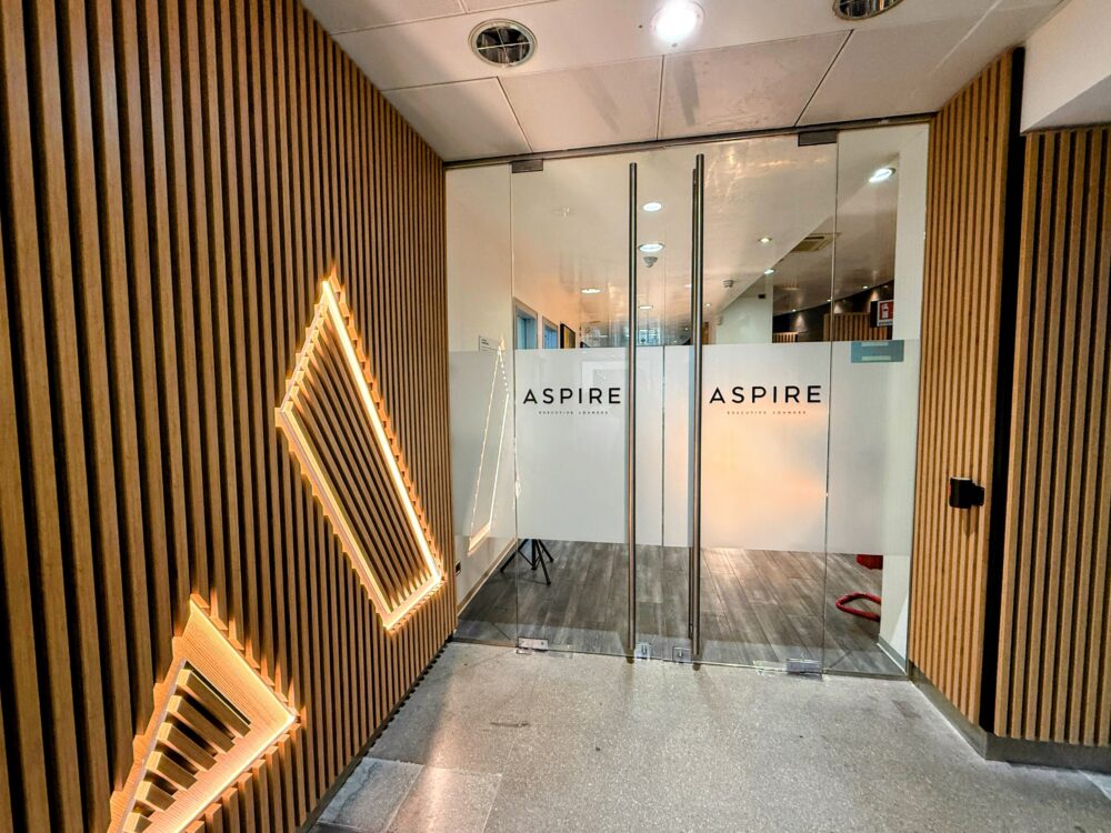 木格子の壁と三角形の照明装飾が特徴的な、Aspire Executive Loungeのガラス張りの自動ドアエントランス。