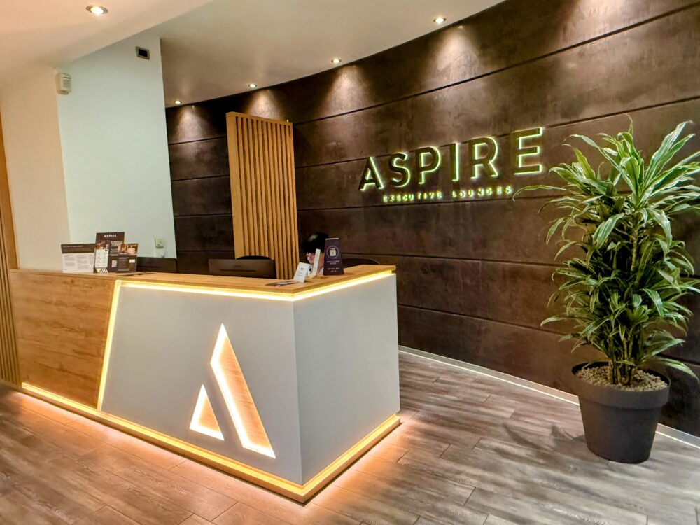 「ASPIRE EXECUTIVE LOUNGES」の立体ロゴが掲げられたレセプションカウンターと、温かみのある木目調のインテリア。
