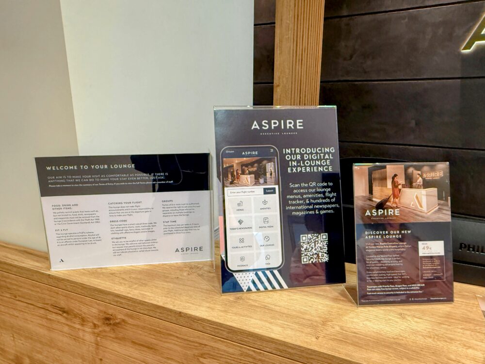 Aspire Executive Loungeの受付に置かれた利用規約のプレートと、入場料49ユーロ・営業時間8:00〜20:00・プライオリティパス無料利用可・ファストトラック込みを記した案内カード。