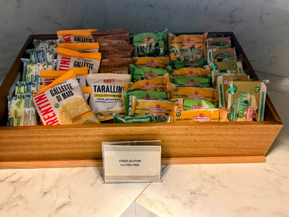 「SENZA GLUTINE(GLUTEN FREE)」の案内プレートが置かれた、グルテンフリーのクラッカーやビスケットなどの個包装スナックが並ぶ木製トレイ。