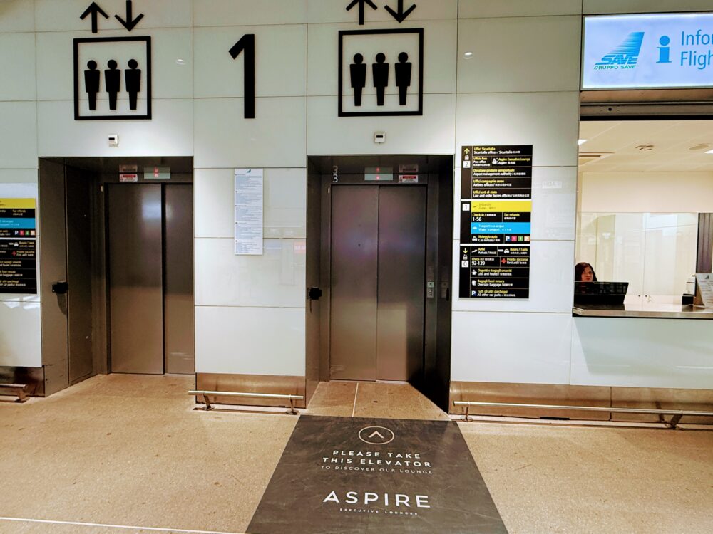 「PLEASE TAKE THIS ELEVATOR TO DISCOVER OUR LOUNGE ASPIRE」と書かれたフロアマットが敷かれた、ラウンジへ向かうエレベーターホール。