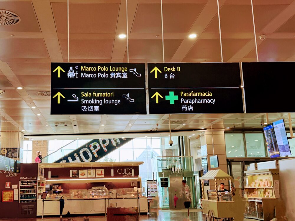 「Marco Polo Lounge」や「Smoking lounge」の方向を示す上部の黒い案内標識と、下のショップエリアを見下ろした景色。