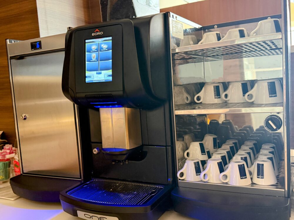 青い照明でライトアップされたEGRO製の全自動コーヒーマシンと、綺麗に棚に収納されたLAVAZZAロゴ入りの白いコーヒーカップのセット。