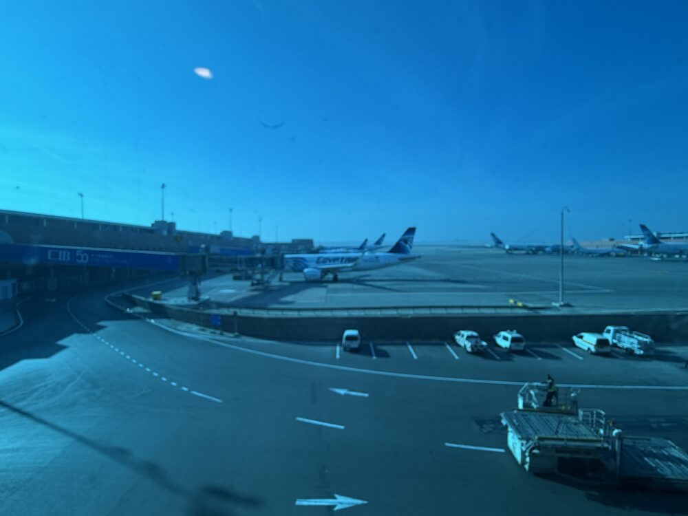 丸窓のガラス越しに広がる、カイロ空港の明るい滑走路と青空の風景。