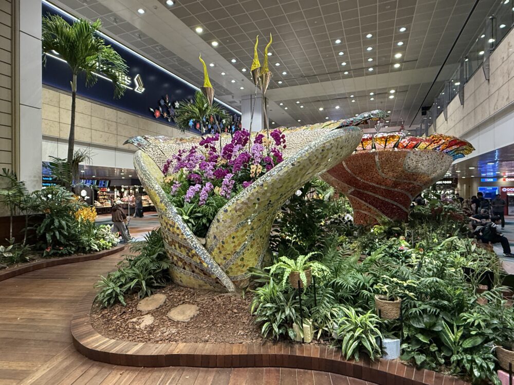 シンガポール・チャンギ空港第2ターミナルの屋内庭園。黄色とオレンジのモザイクタイルで装飾された大きな花器に紫と白の蘭が咲き、奥にシンガポール航空の看板が見える