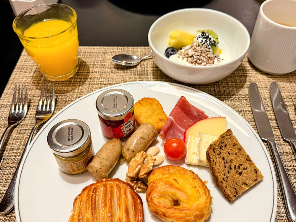 カトラリーやドリンク、ボウルに入ったヨーグルト、パン、ハム、チーズなどが並ぶ朝食の卓上風景