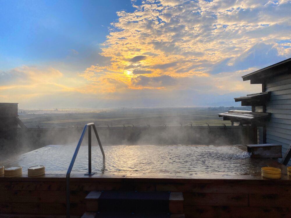 熊本県の露天風呂から見る朝焼け。湯けむりの向こうに平野と山並みが広がる。