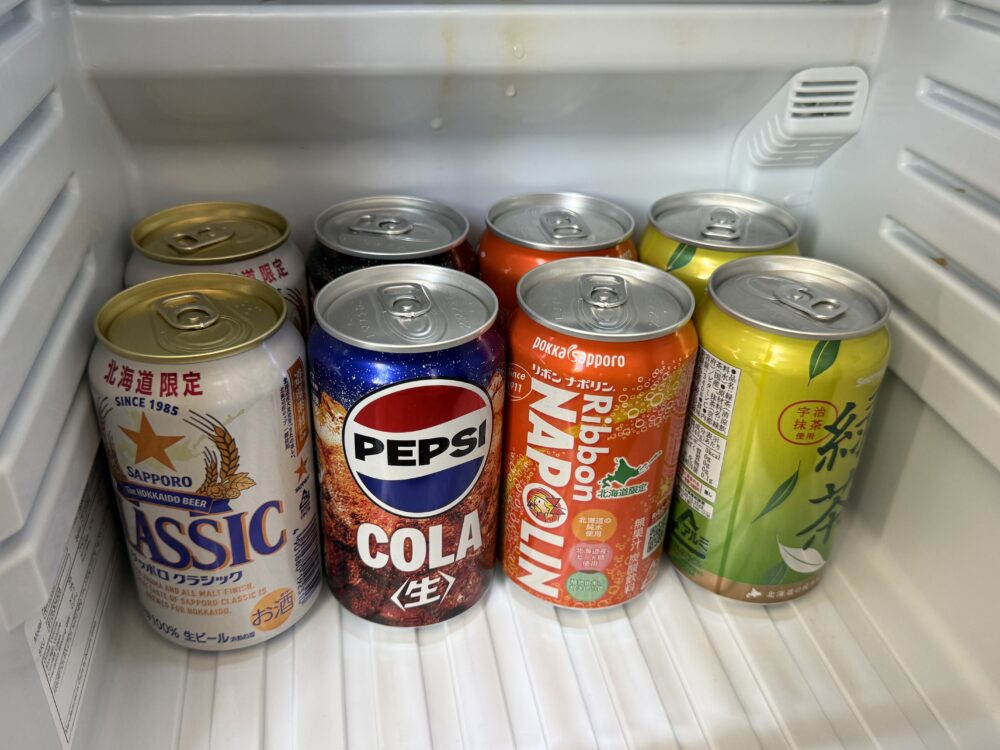冷蔵庫の中に並ぶドリンクのアップ。サッポロクラシック、ペプシコーラ、リボンナポリン、伊藤園の緑茶などの缶が整然と並んでいる。