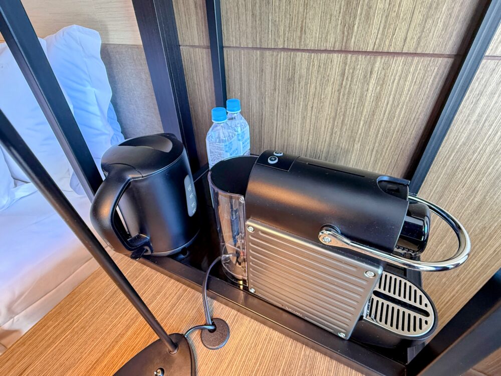 客室の棚に置かれた黒い電気ケトルとカプセル式のネスプレッソコーヒーメーカー、およびミネラルウォーターのボトル。