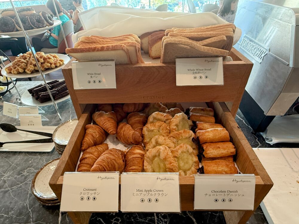 朝食のベーカリーコーナー。木製の棚に食パン、クロワッサン、ミニアップルクラウン、チョコレートデニッシュなどが種類豊富に並んでいる。