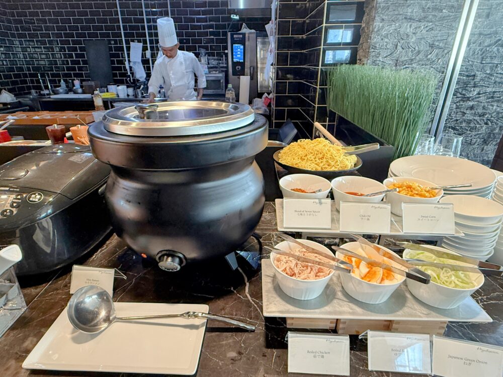 東山ニセコビレッジ・リッツ・カールトン・リザーブの朝食ビュッフェにあるラーメンステーション。大きな黒い寸胴鍋の横に、茹で上げられた麺と、鶏肉、半熟卵、ネギなどの薬味、そしてお玉が用意されている。