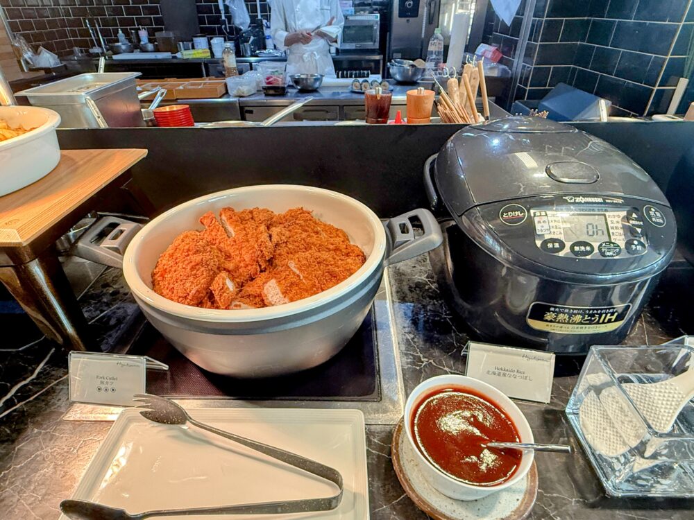 朝食ビュッフェのホットミールコーナー。大きな白い鍋に揚げたての豚カツが山盛りにされており、横には自動炊飯器と、味噌カツのソースが入った白いボウルが置かれている。