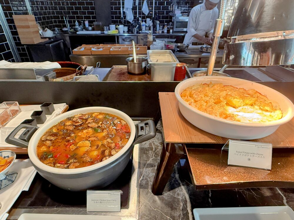 朝食ビュッフェの温かい料理。大きな鍋に入った具だくさんの「Japanese Chicken Soup Curry(日本の鶏スープカレー)」と、グラタン皿で焼かれた「Baked Potato with Cheese(ベイクドポテトチーズ入り)」が並ぶ。