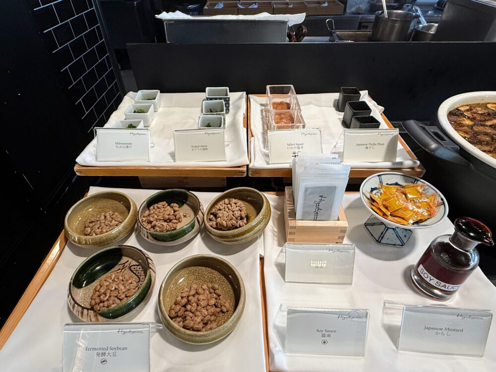 朝食ビュッフェの和食コーナー。手前には納豆の入った陶器の器が複数並び、奥には「Mibunazuke(壬生菜漬け)」、「Pickled Okura(オクラの漬物)」、「Salted Squid(いかの塩辛)」、「Japanese Pickle Plum(梅干し)」などの小鉢が提供されている。