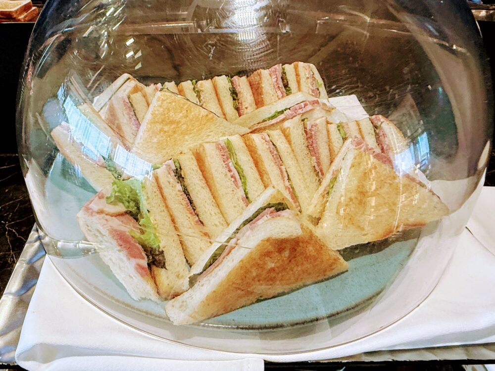 朝食ビュッフェのサンドイッチ。トーストされたパンにハムとレタスが挟まれたサンドイッチが、透明なドーム型のカバーの中に綺麗に並べられている。
