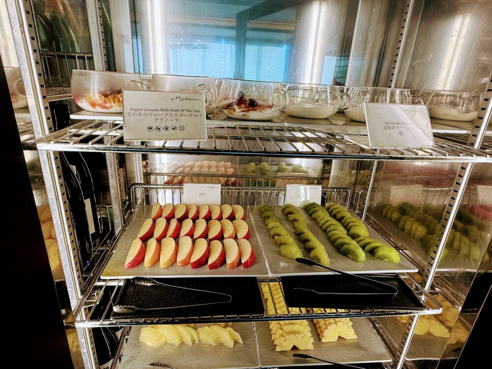 朝食ビュッフェの冷蔵ショーケース。上段にはヨーグルトやフルーツ入りのグラスデザート、中段にはカットされたリンゴとキウイ、下段にはパイナップルが美しく冷やされている。