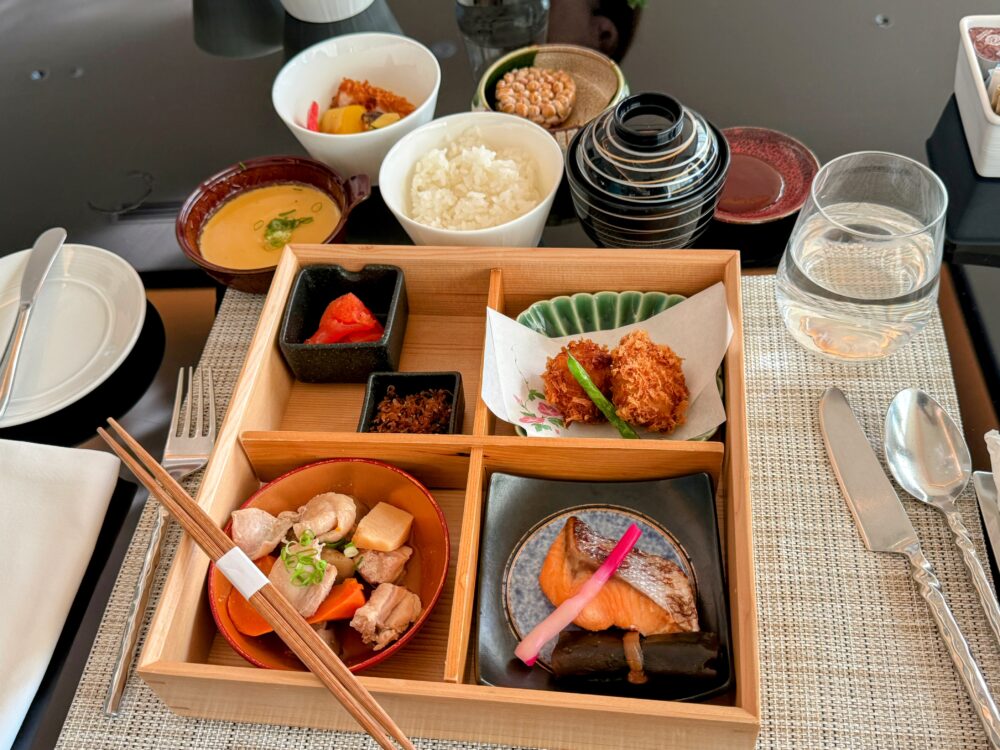 東山ニセコビレッジ・リッツ・カールトン・リザーブの朝食。木製の弁当箱のような和朝食セット(煮物、焼き魚、明太子、揚げ物)を中心に、ご飯、味噌汁、納豆、ジュース、水が並んでいる。箸も添えられ、上品な和定食の雰囲気が伝わる。